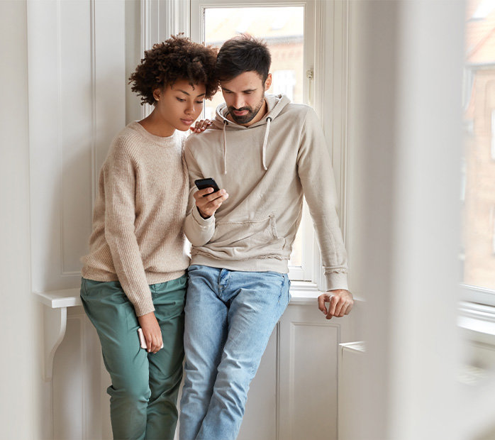 Couple leaning against apartment window looking at phone together.